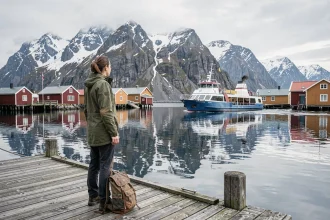 Voyageuse avec sac à dos attendant le ferry aux îles Lofoten, chalets de pêcheurs rouges et montagnes enneigées en arrière-plan